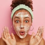 Close up portrait of astonished woman washes face after shower, has soap bubbles on complexion, rais