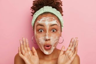 Close up portrait of astonished woman washes face after shower, has soap bubbles on complexion, rais