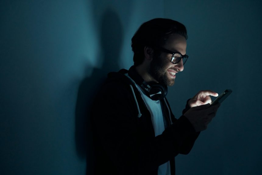 Confident young man smiling while using smart phone in the dark