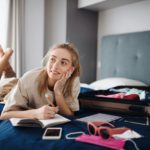 Young woman writing notes and packing clothes into suitcase indoors at home, planning holiday.